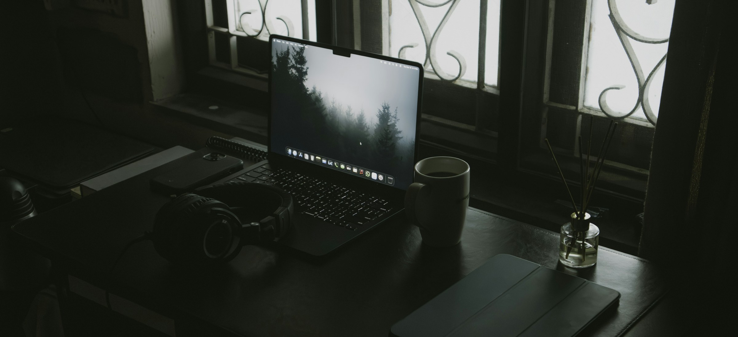 A laptop, headphones and a coffee cup on a desk by a window — remote work setup